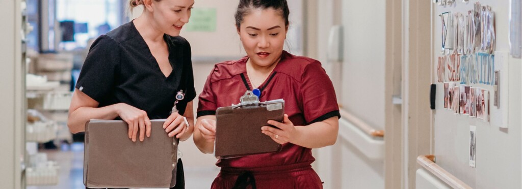 Photo of two female healthcare professionals in scrubs looking at a clipboard in a hospital setting.