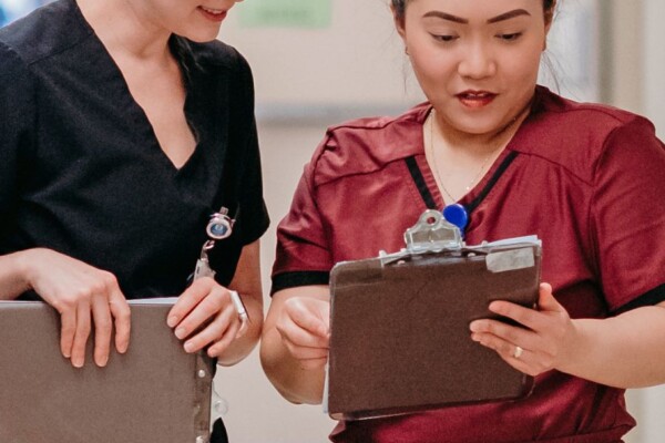 Photo of two female healthcare professionals in scrubs looking at a clipboard in a hospital setting.