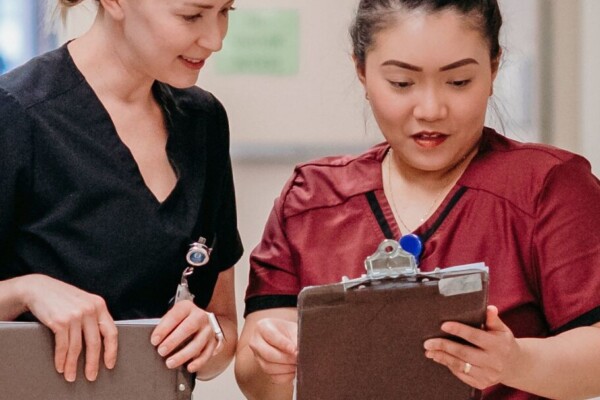 Photo of two female healthcare professionals in scrubs looking at a clipboard in a hospital setting.