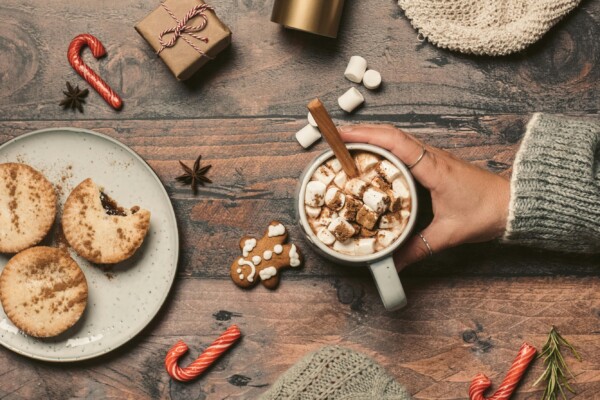 Photo of hand holding mug of hot chocolate surrounded by candy canes, cookies, and a wrapped present.