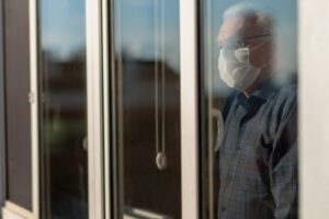 An elderly man peers out of a window while wearing a mask.